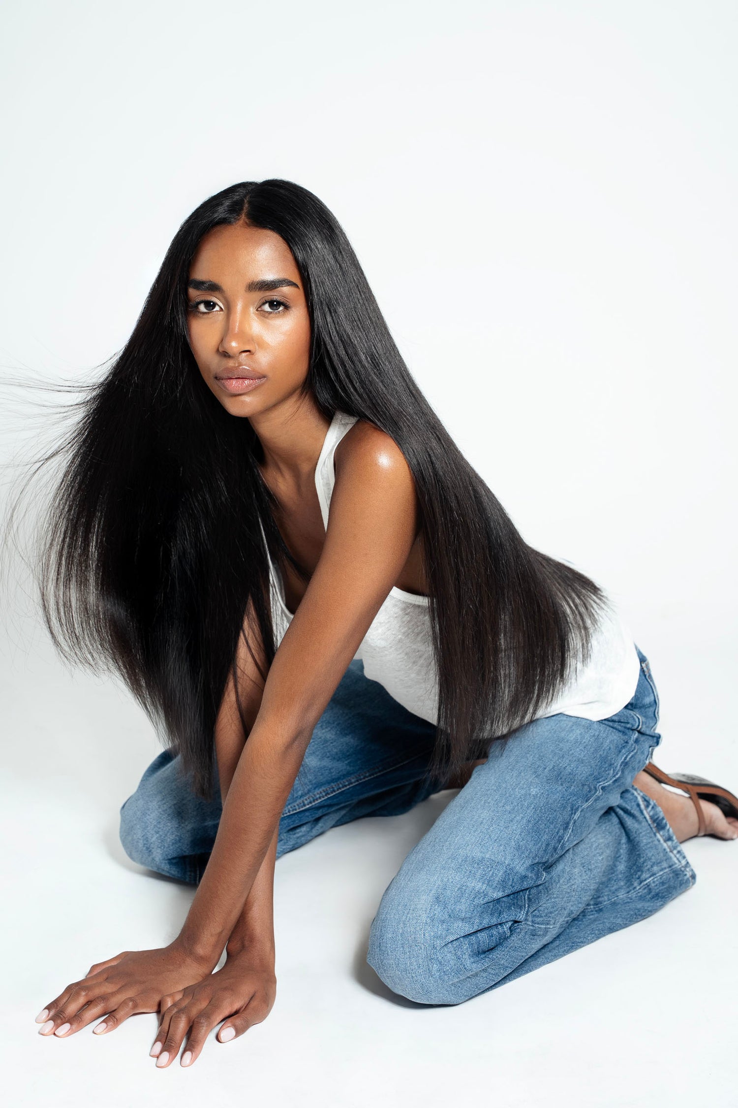 Woman with long, straight black hair wearing a white tank top and blue jeans on a white background