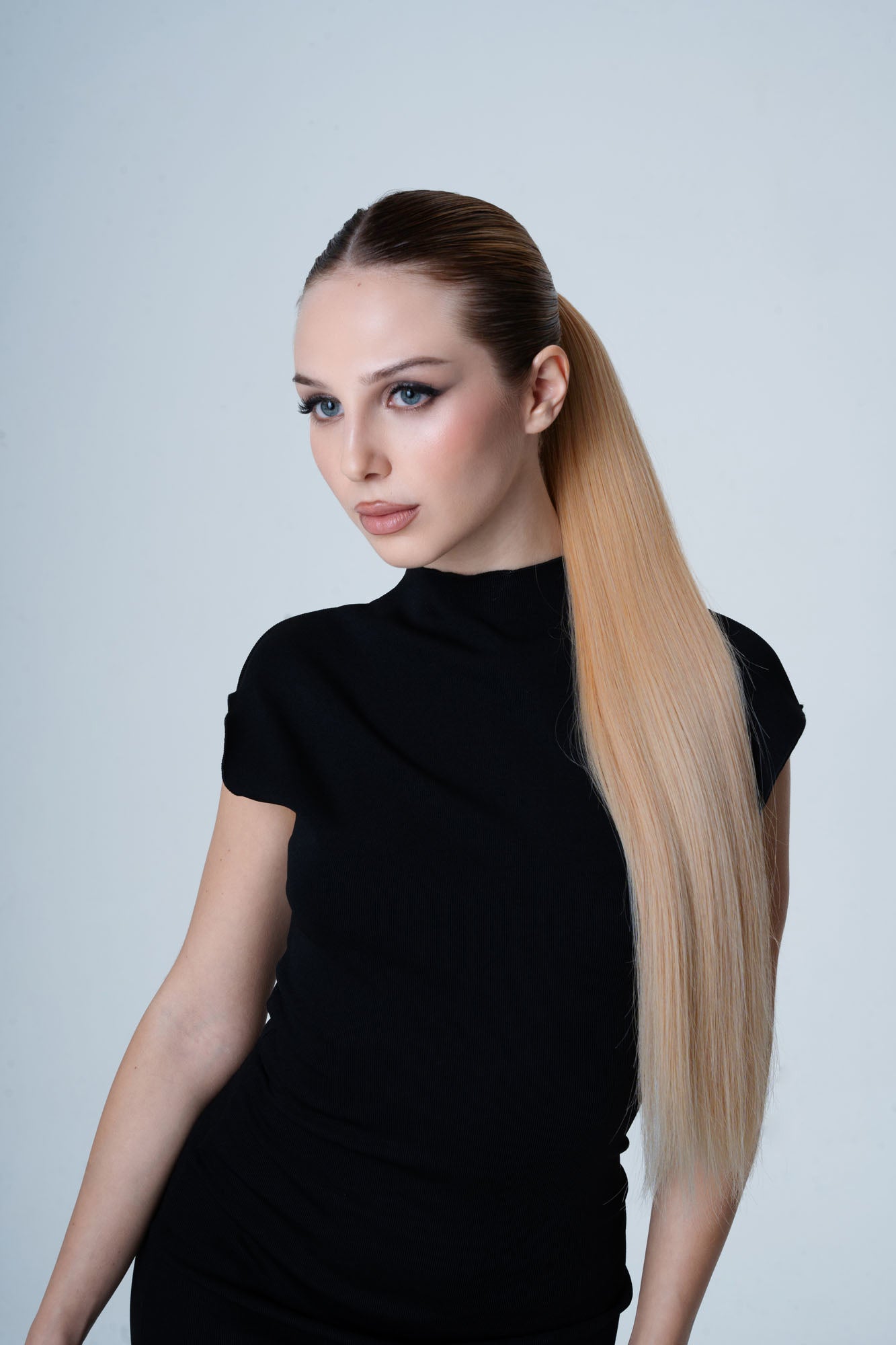Woman with long blonde hair styled in a high ponytail by 1010 Extensions against a light gray background
